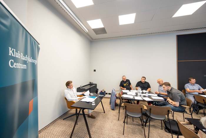 Szeroki kadr sali konferencyjnej: uczestnicy siedzą przy zestawionych stołach.