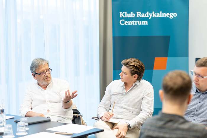 Dwie osoby rozmawiają, jedna wyjaśnia coś ruchem rąk; w tle roll-up klubu.